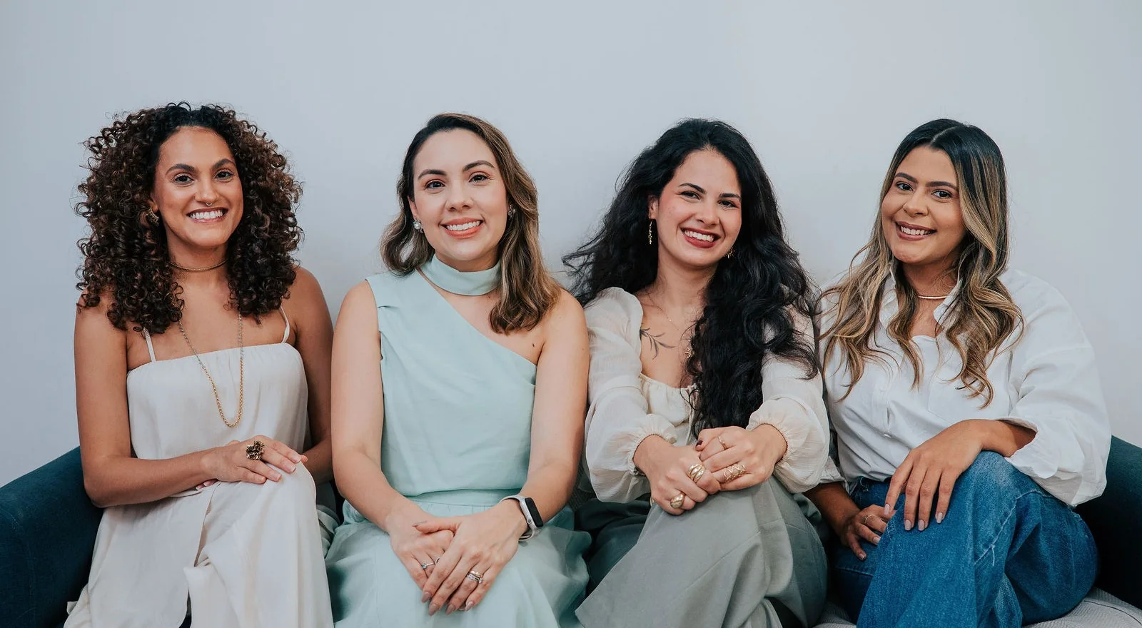 Dharana Vieira, Brenna Damasceno, Milena Coelho e Maria Angélica Lacerda sentadas juntas num sofá, sorrindo